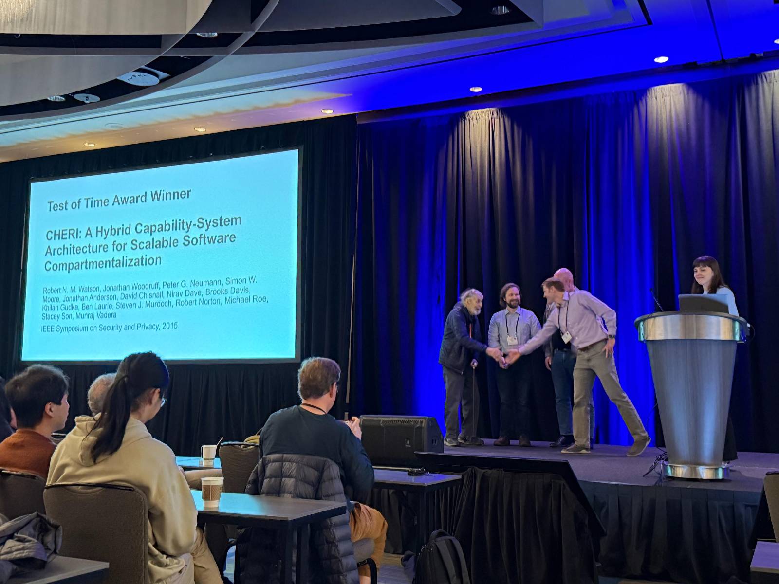 Peter G Neumann shaking hands with the presenter while receiving the IEEE Security and Privacy Test of Time Award, with some of the other coauthors on stage.  A title slide shows the paper title and complete author list